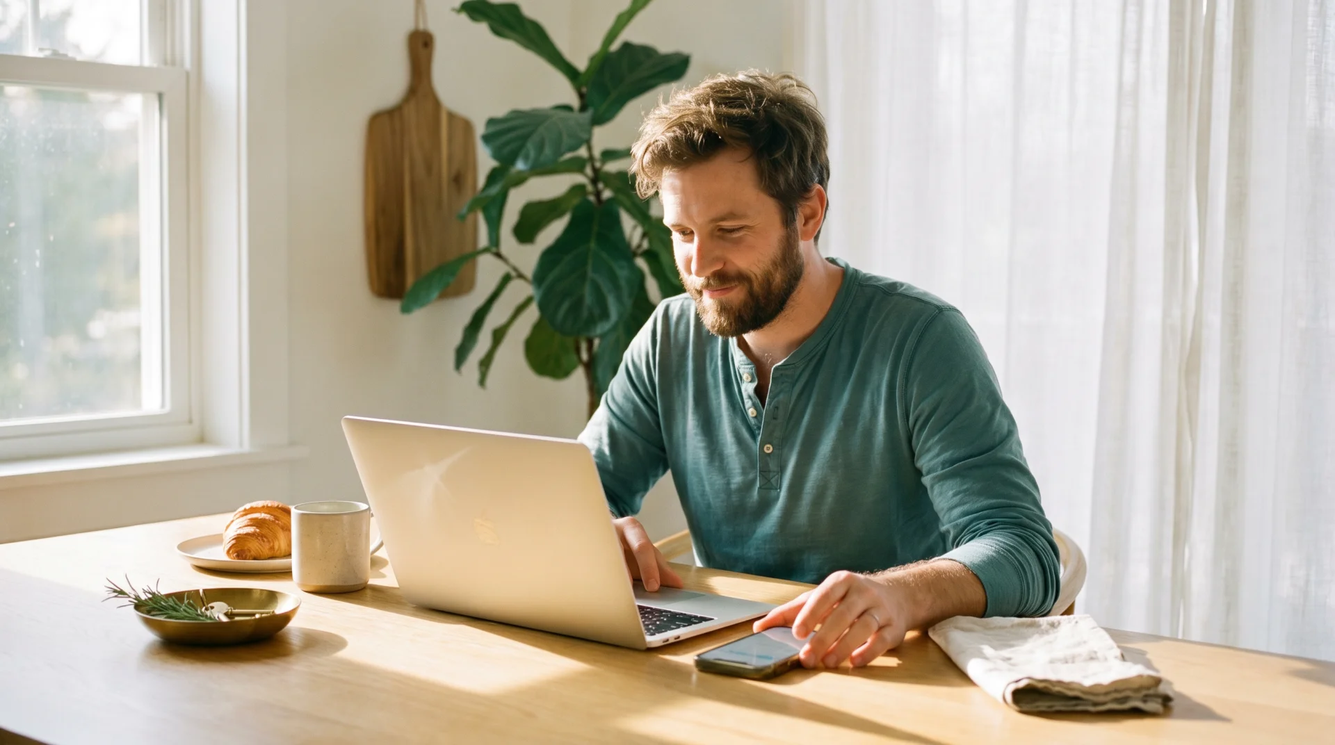 Solo operator at a sunlit kitchen table, laptop open, phone face-up next to him — moving fluidly between clicking and asking